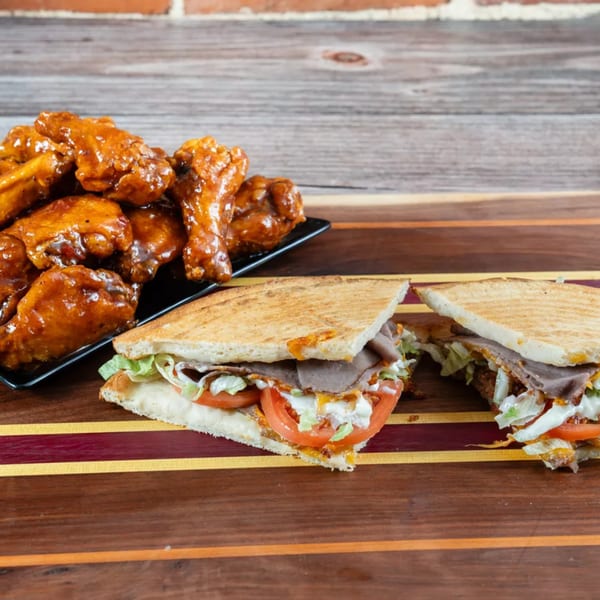 Punxsy Pizza's grilled roast beef sandwich with lettuce, tomato, and sauce, next to a plate of chicken wings.