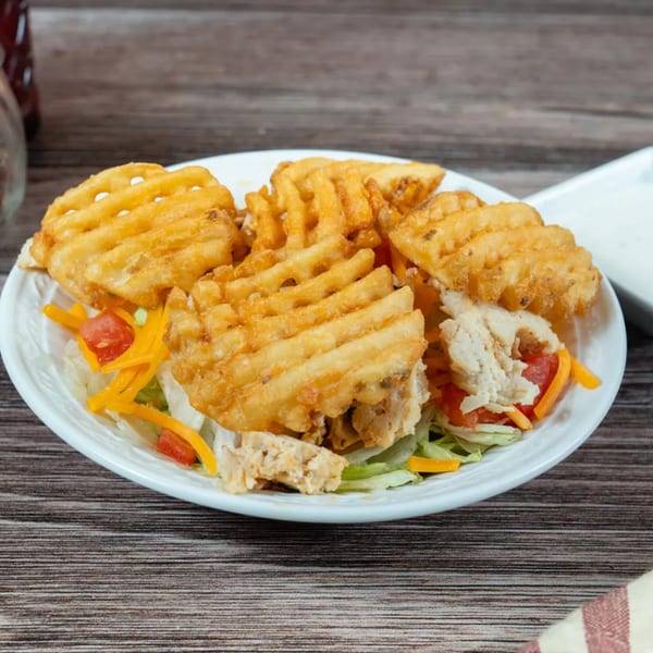 A white plate of waffle fries, chicken, lettuce, tomatoes, and shredded cheese on a wooden table.